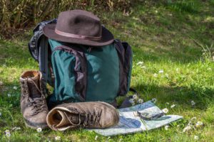 Auf dem Bild sieht man einen Wanderrucksack mit Wanderhut und Schuhen, die im Gras stehen. Davor liegt eine Wanderkarte mit einem Kompass darauf.